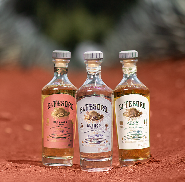 Three bottles of El Tesoro tequila side by side on the mineral-rich soil of the La Alteña distillery. 
